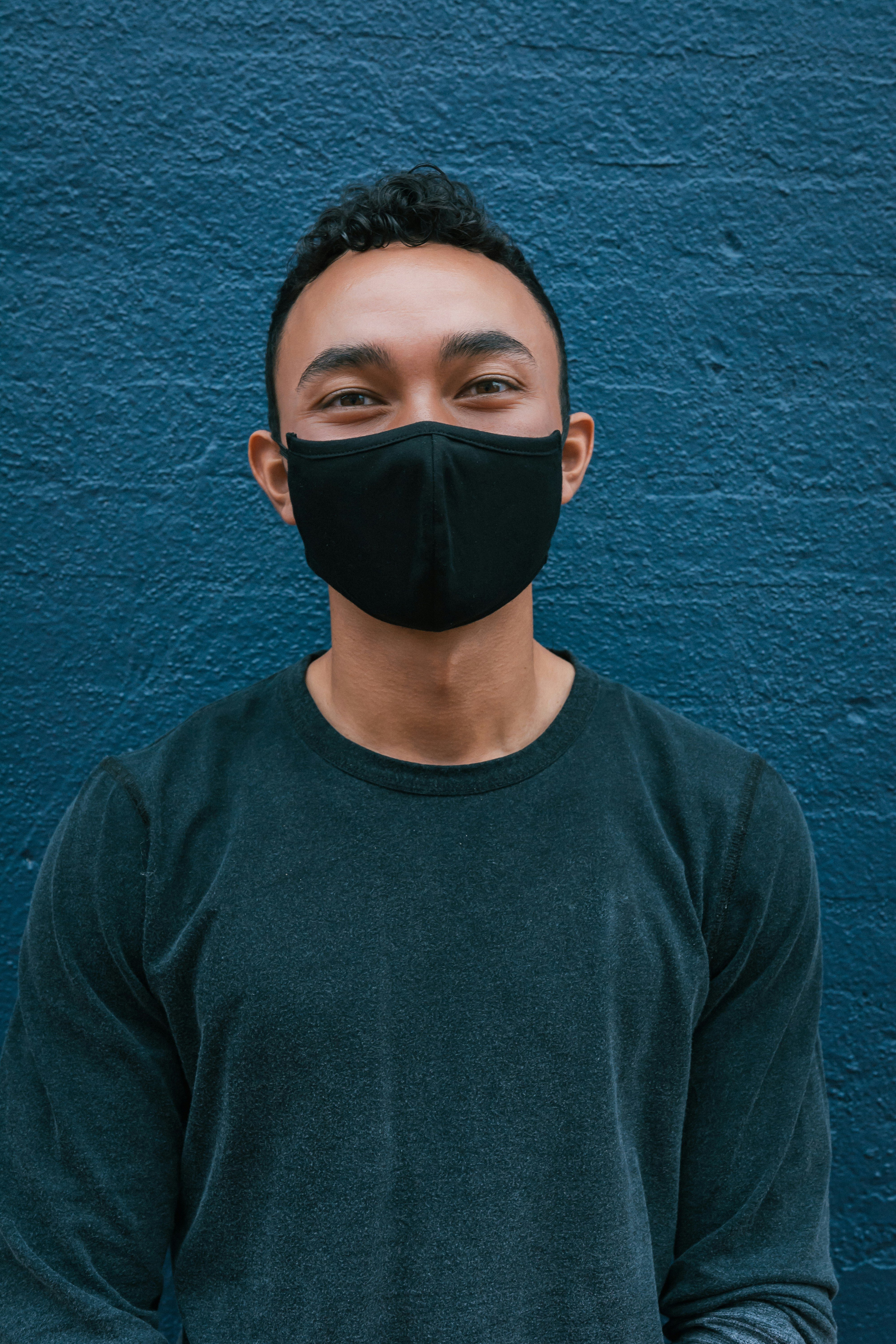 A young man stands in front of a blue brick wall wearing a dark blue shirt and a black facemask