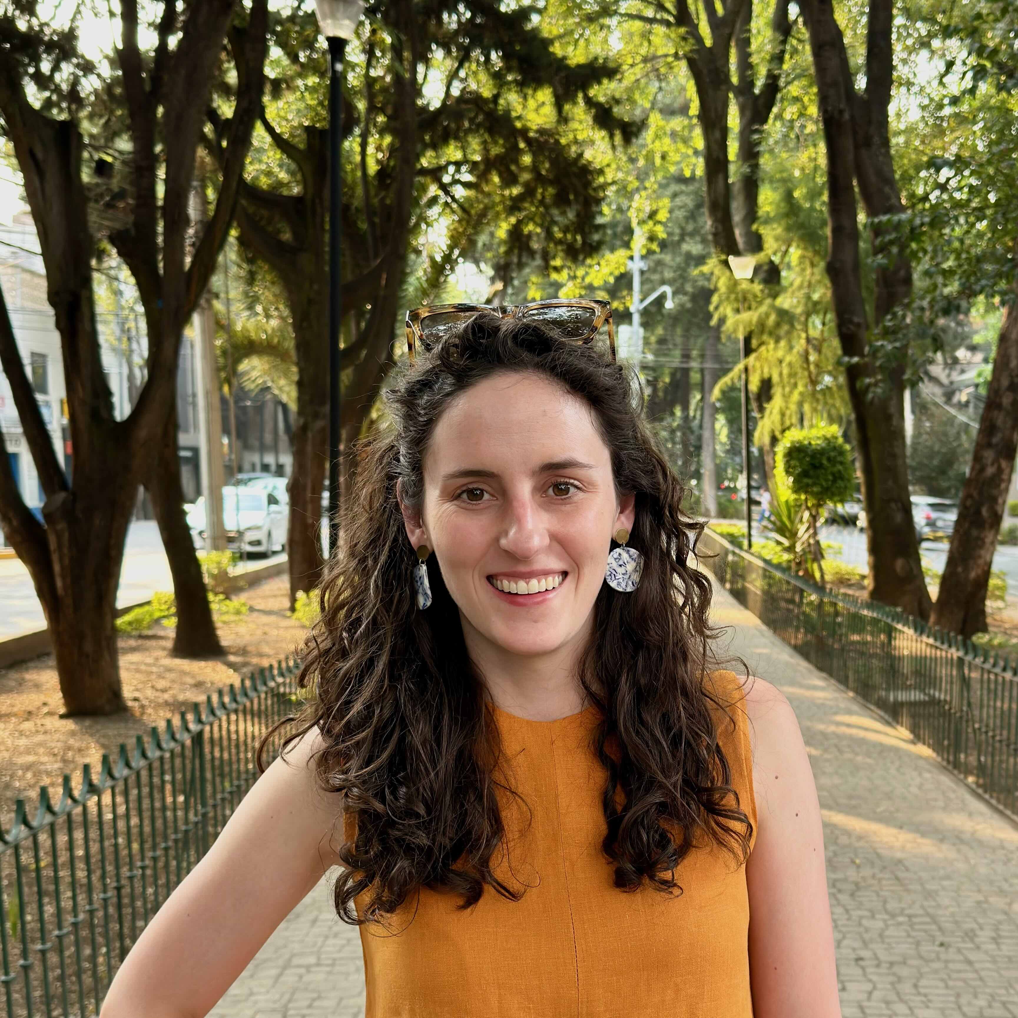 Headshot of the site author. Author has brown curly hair, is standing in front of a path lined with trees and is looking straight ahead and smiling.
